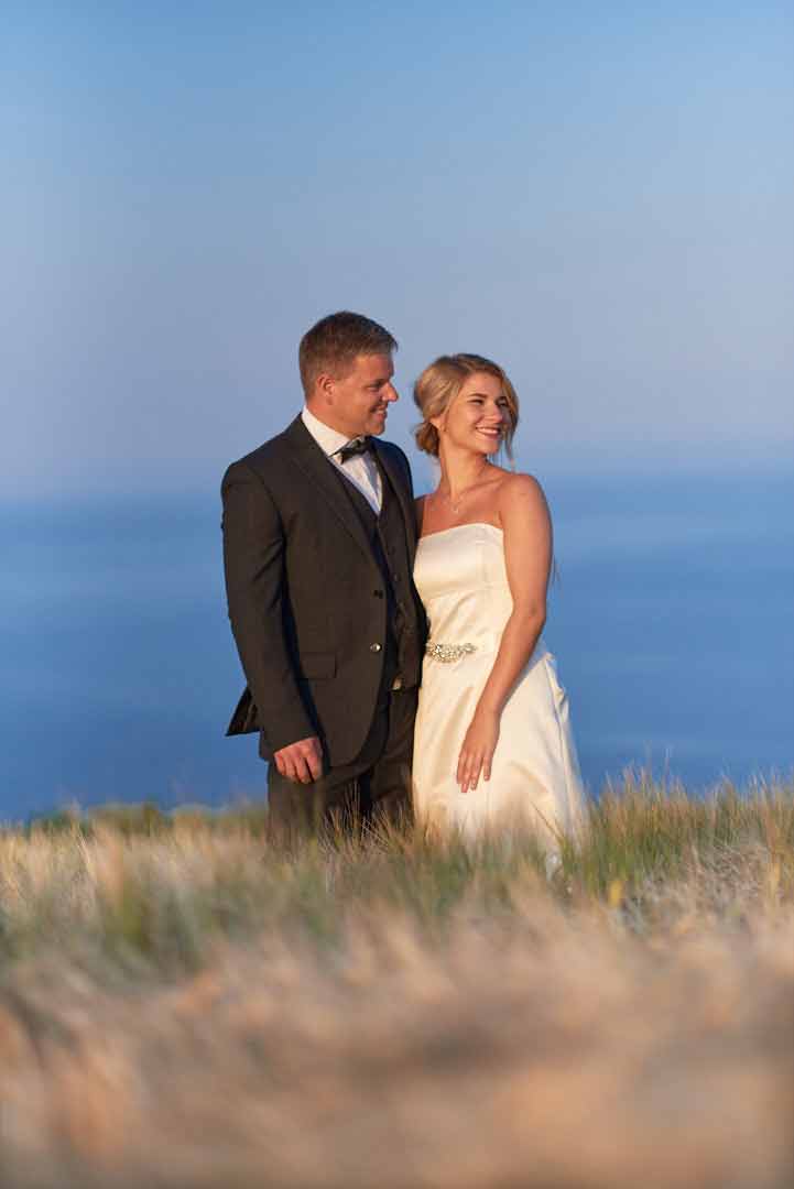 Full body portrait of bride and groom standing in long grass with blue sea in the background. They are standing close and looking off into the distance and bot are smiling