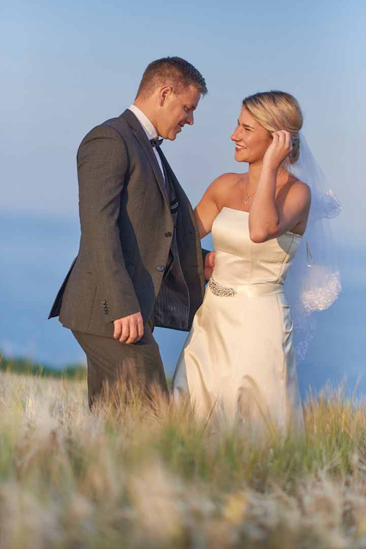 Full body portrait of bride and groom standing in long grass up to their knees. They are standing close and about to embrace.