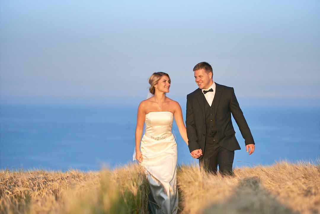 Bride and groom walk towards camera, thier eyes lock and they smaile at each other. The blue sea behind them merges with the bright blue sky