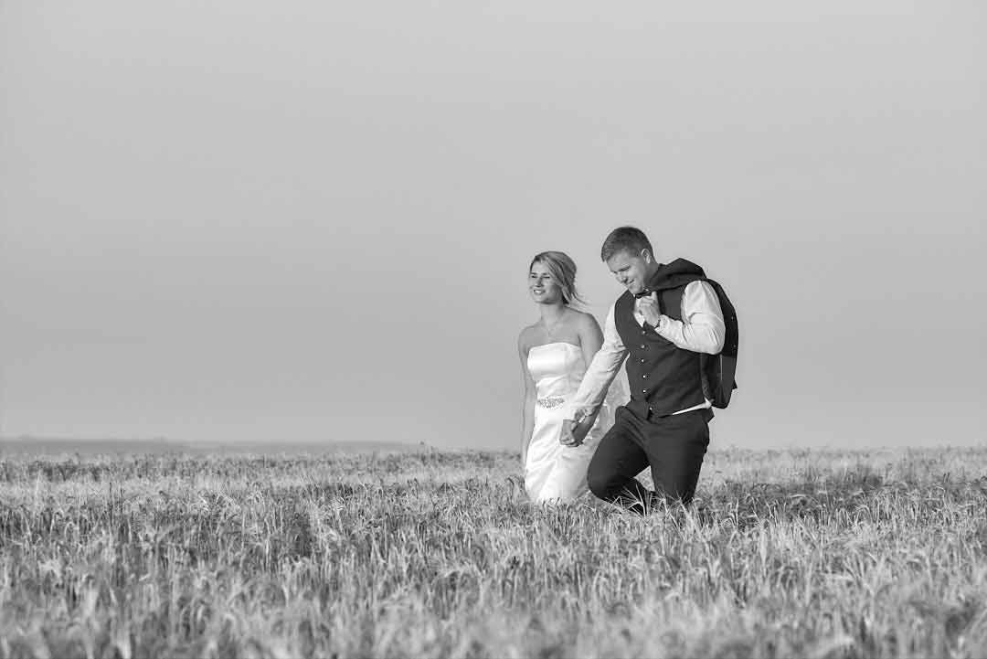 A bride and groom walk hand in hand through a hay field with a serene, hazy sky in the background. The bride wears a strapless gown, and the groom is holding his black jacket over his left shoulder.