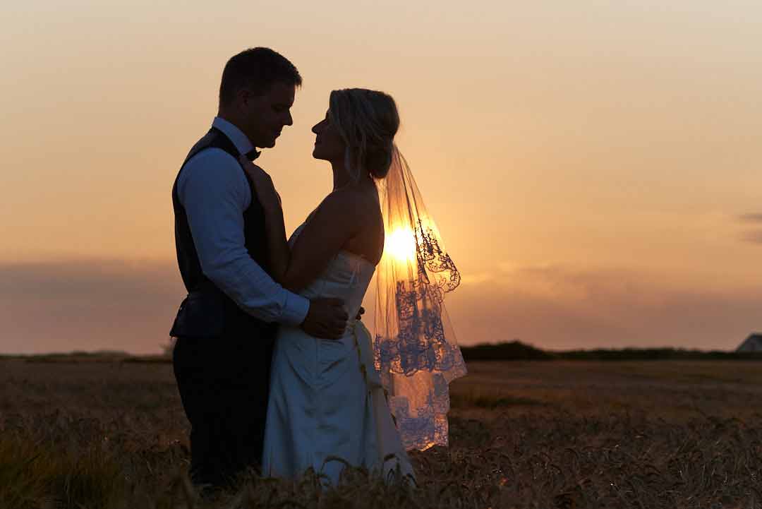 Silhouette of a bride and groom standing in a field. The big yellow sun is setting in the background catching the brides veil