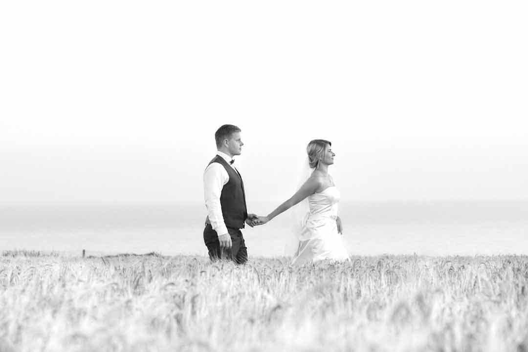 A bride and groom walk hand in hand through a field with a serene, hazy sky in the background. The bride wears a strapless gown, and the groom a suit vest and bow tie.