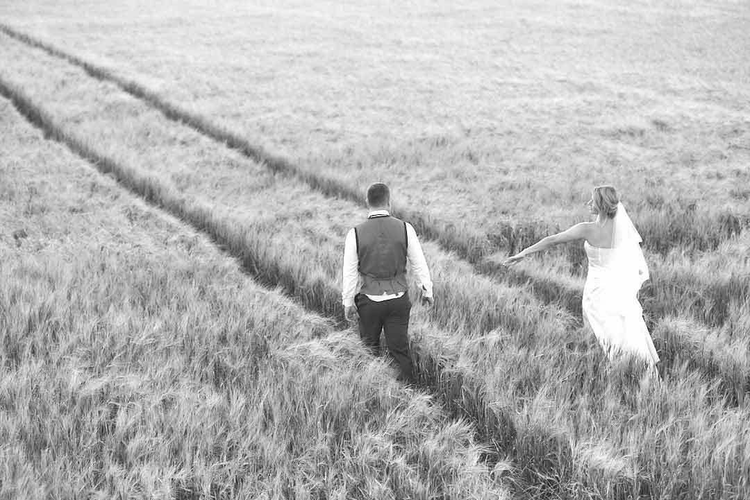 Bride and groom walking in a hay field with two tracks that look like train tracks leading off into the distance. The bride extends her left hand hoping the groom will hold her hand