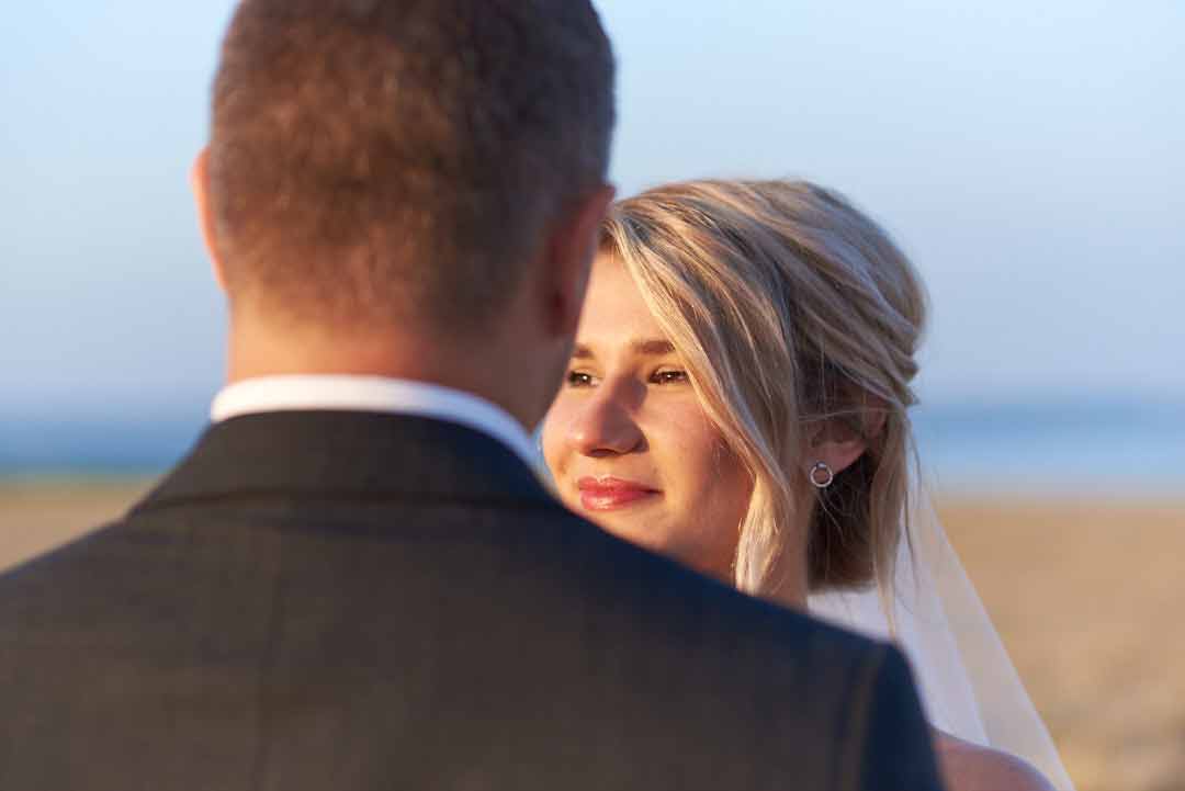 Close up of a bride's face taken from behind her grooms head. Beautiful soft yellow evening sunlight illuminates the brides face