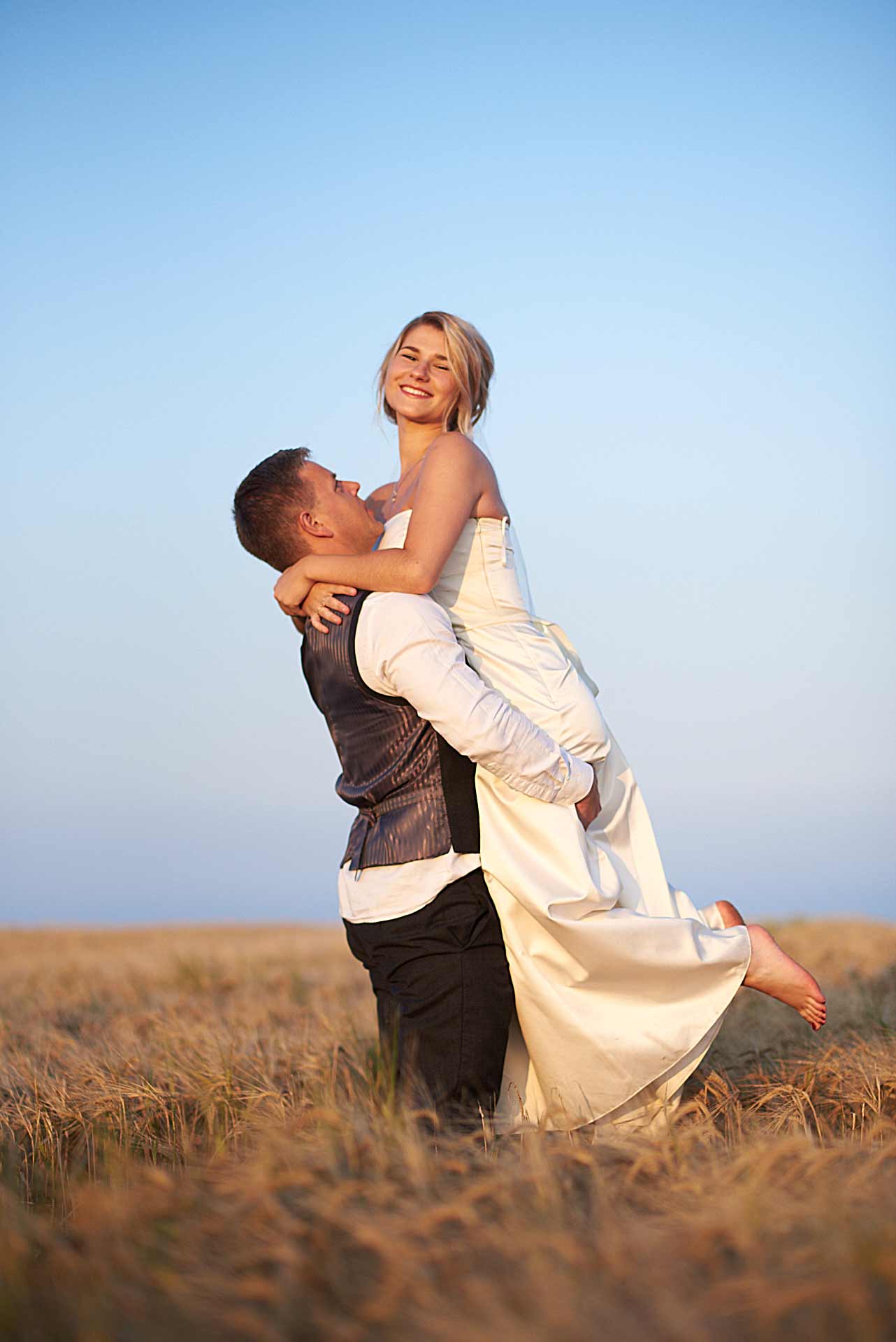 Newlyweds stroll along coastal cliffs, bride in white, groom in suit, hand in hand, smiling, surrounded by scenic beauty.