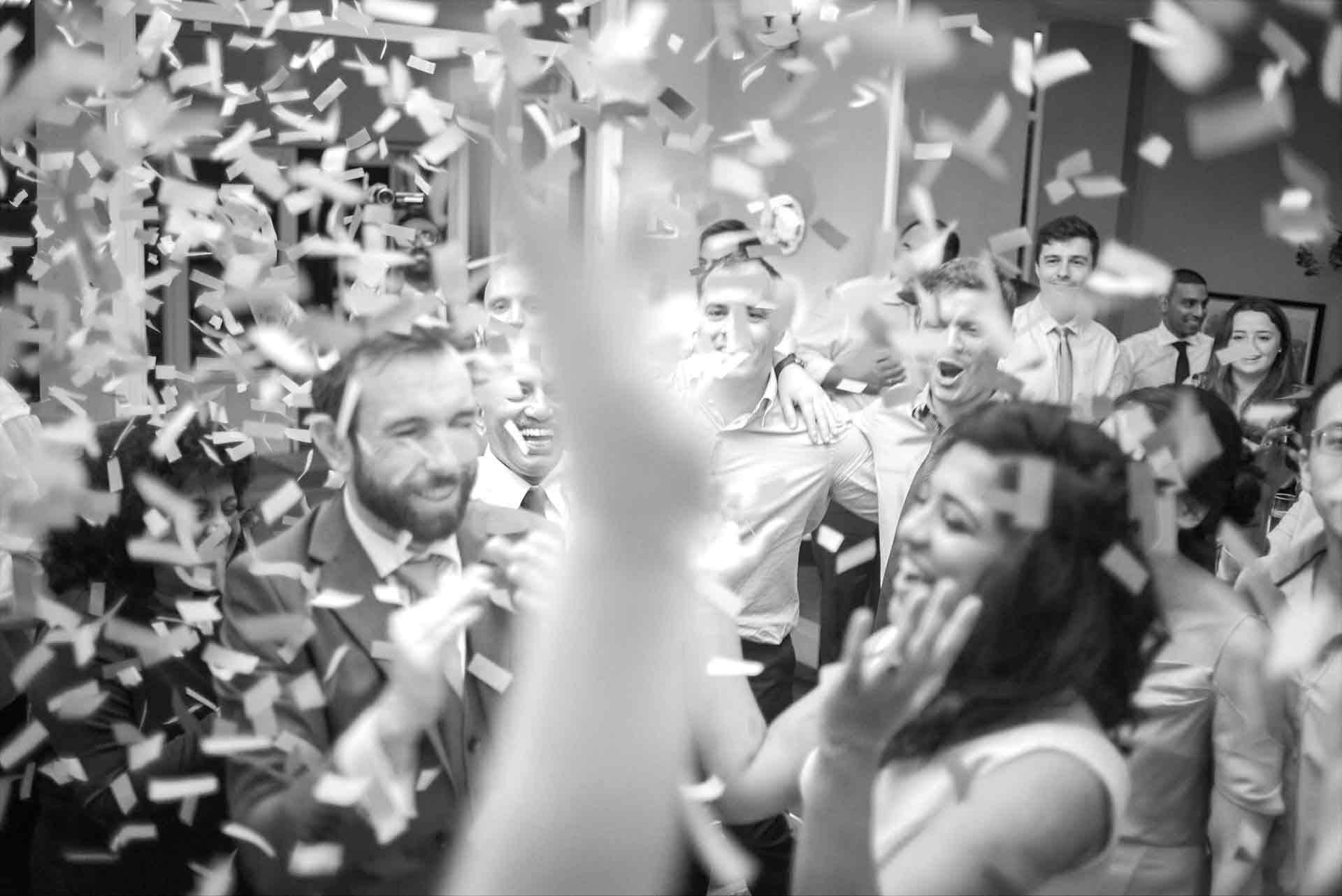 Confetti rains from the sky after the wedding couple's first dance in the hotel. Wedding guest look on enjoying themselves while an arm is raised in front of the camera