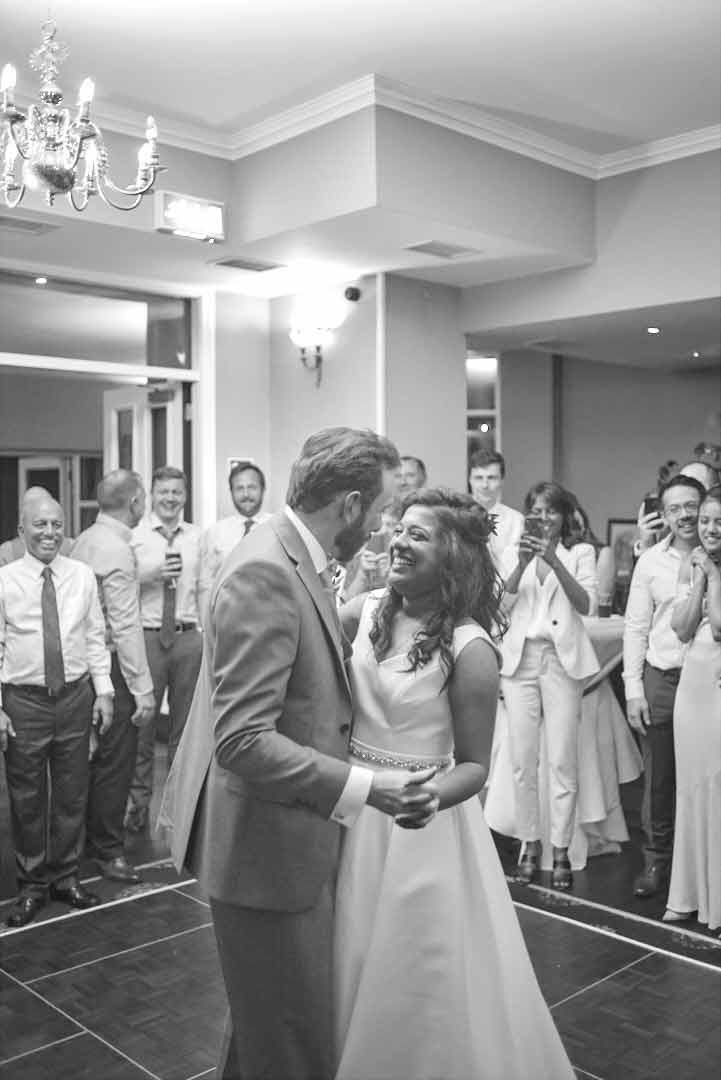 Wedding couple enjoying their first dance while the wedding guests stand behind them looking on. Some record the moment with their mobile phones.