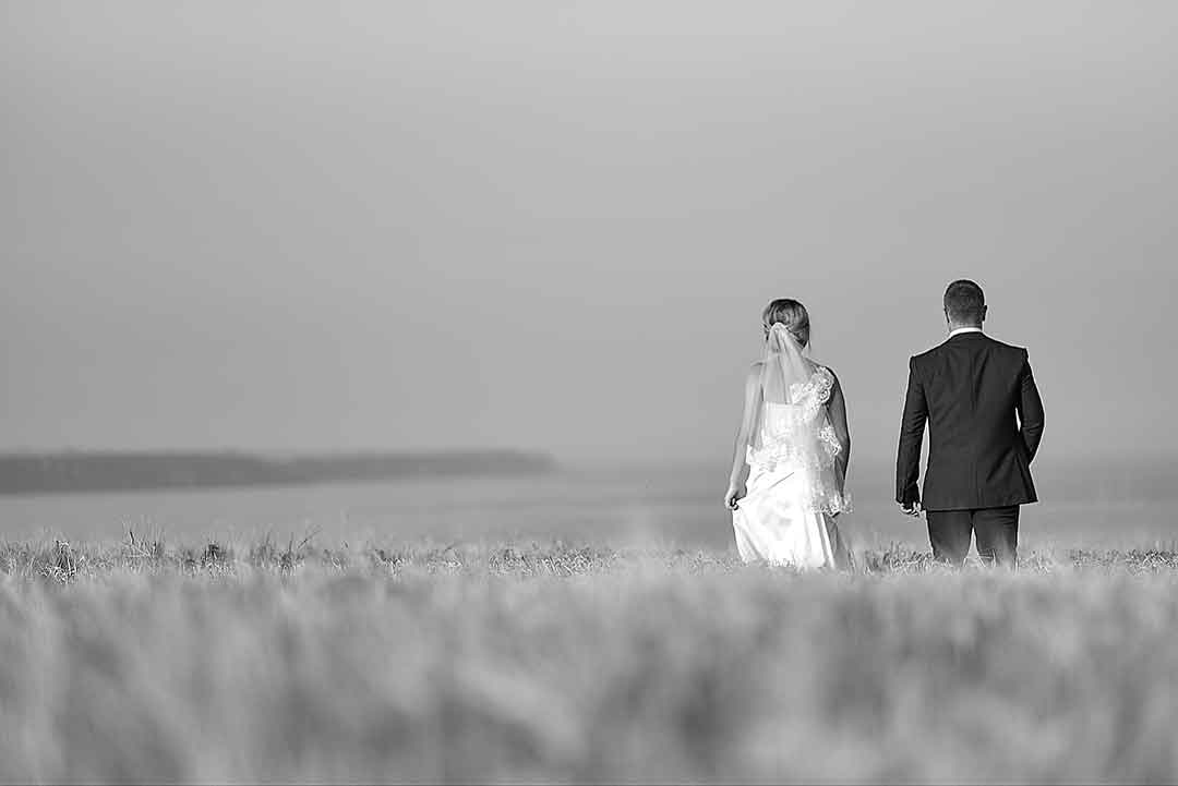 A bride and groom walk hand in hand through a hay field with a serene, hazy sky in the background. The bride wears a strapless gown, and the groom is holding his black jacket over his left shoulder.