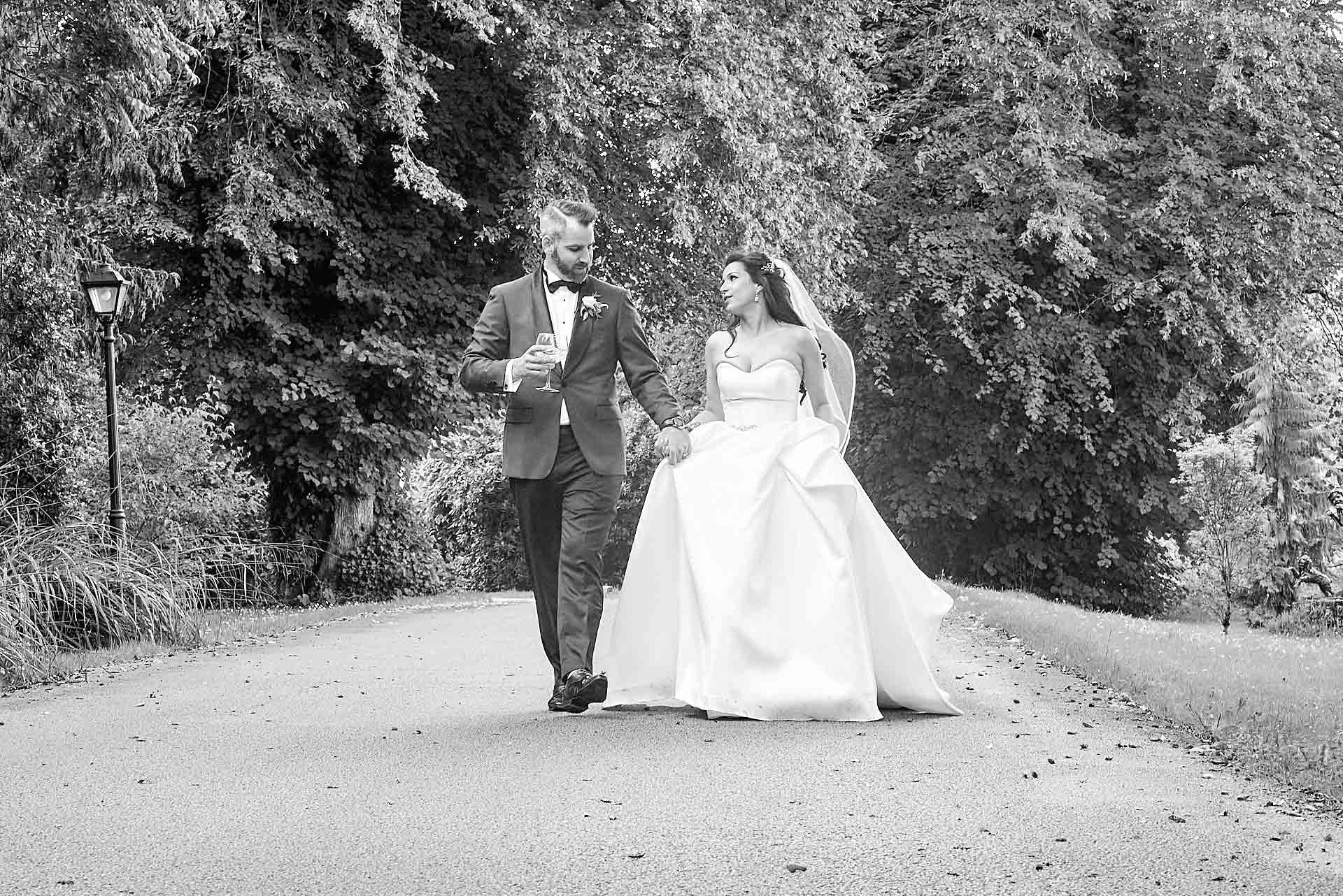 Black and white shot of a bride and groom walking toward the camera on a big gravel path. They are holding hands, deep in conversation. Background is covered in thick trees