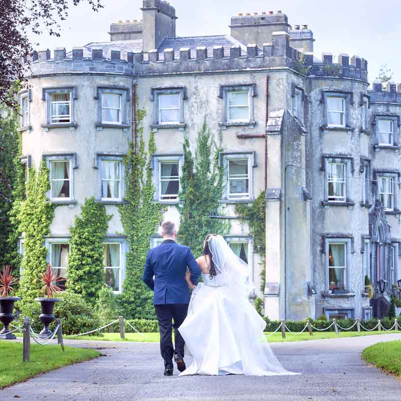 Bride and groom walk arm in arm down a path towards Ballyseede Castle Hotel